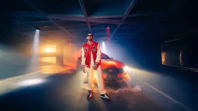 Artista de pie con chaqueta roja frente a un auto rojo en un garaje con haces de luz azul y anaranjada