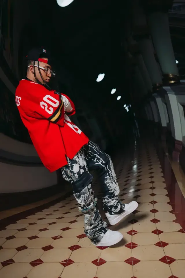 Artista en pose inclinada con camiseta roja, pantalón camuflado y tenis blancos, en un pasillo de baldosas rojas y blancas