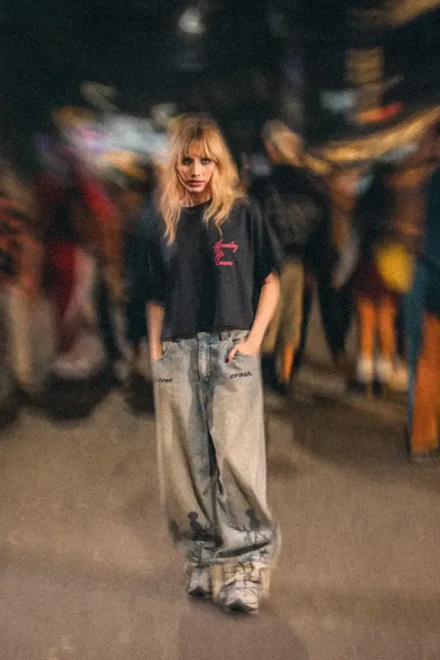 Mujer rubia con camiseta oscura y jeans amplios de pie frente a un fondo de personas en movimiento desenfocado