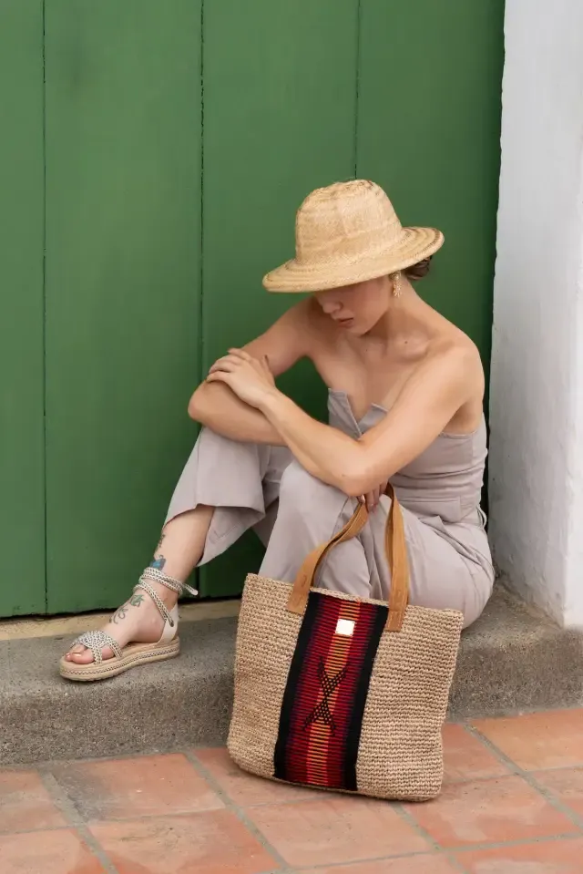 Mujer con sombrero de paja sentada frente a una puerta verde, con un bolso tejido a su lado