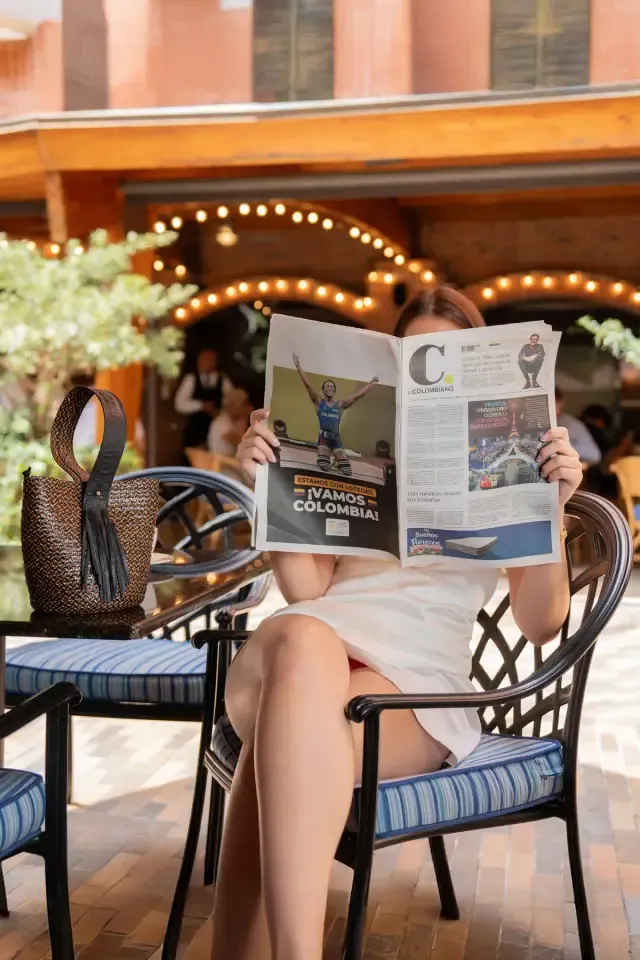 Mujer leyendo un periódico en una terraza con luces cálidas, con un bolso de mimbre sobre la silla