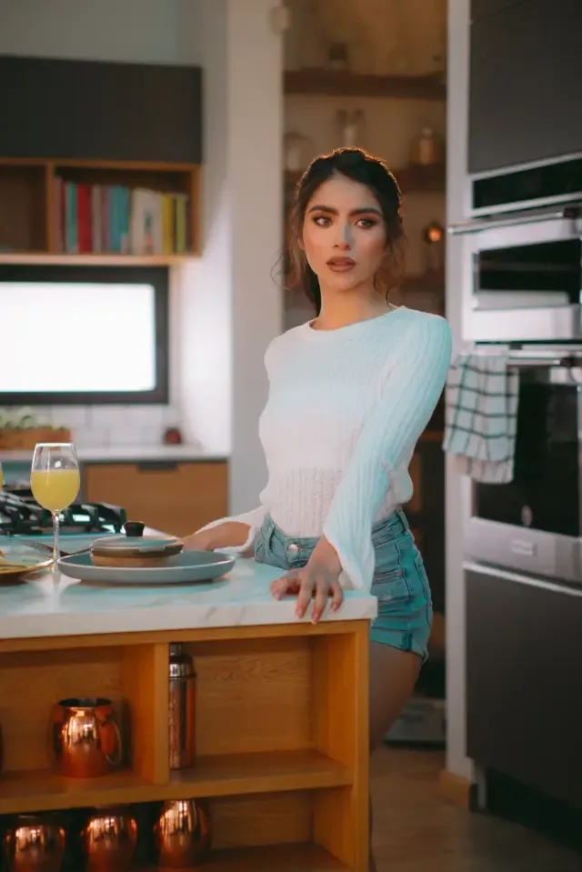 Mujer apoyada en la isla de una cocina luminosa, con un jugo y un desayuno servidos