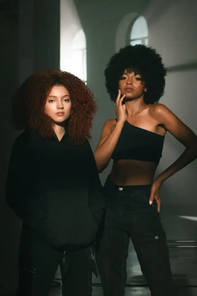 Dos modelos con ropa negra y cabello rizado posan sobre un fondo oscuro